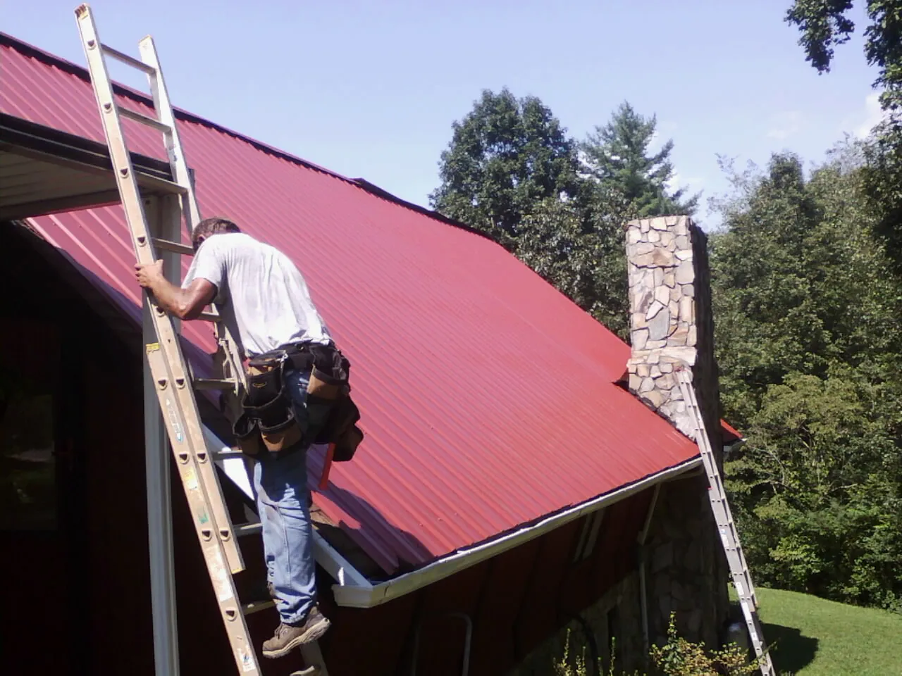 Roofer on a ladder beside a red metal roof and stone chimney — Soffit & Fascia Repair by Roofing Brothers in Lapine